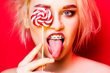 Portrait of a young blonde on a red background. The blonde closes her eyes with candy and shows her...