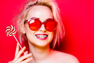 A blonde with short hair smiles with braces on her teeth against a red background. A model in red glasses smiles with candy in her hands. - obrazy, fototapety, plakaty