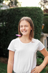 Cute funny smiling playful preteen girl holding pink cosmos flowers in her mouth. Mock up, white shirt. Summer lifestyle, leisure activity, positive emotions