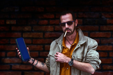 hombre posando con gafas de sol, fumando y con un altavoz inalambrico