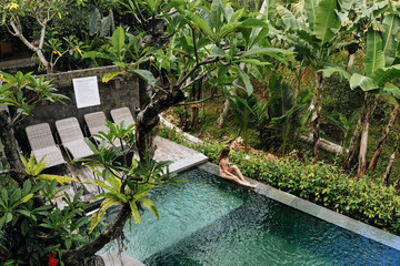 Back view of woman in bikini in a private pool in Bali admires a beautiful view of the palm...