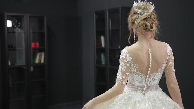 Young blonde bride in white wedding dress with crown on her head is walking forward in room with black walls. Looks over his shoulder at camera smiles