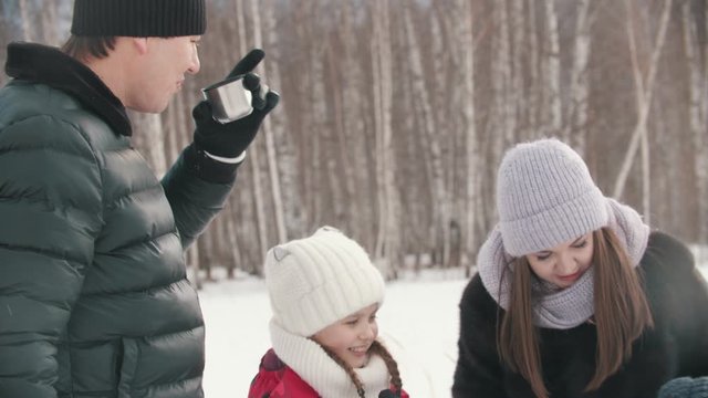 Young family drinking hot drinks from the termos outdoors near the forest