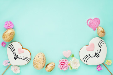 Easter composition on a blue background. Gingerbread cookies with a painted rabbit face, golden eggs, paper flowers and hearts. Flat layout, top view, space for copy..