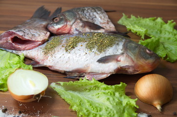 On the table lies a fresh, gutted river fish, crucian carp, onions, lettuce, salt, spices.