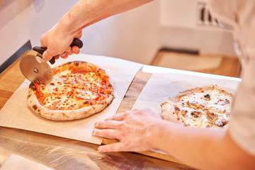 Italian kitchen and cooking concept. Chief with a pizza cutter cutting pizza to pieces at Neapolitan pizzeria.