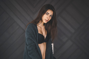 Young brunette in black underwear, knitted knee pads and cardigan sits on a wooden chair. Behind monochrome dark background