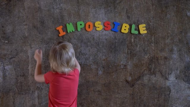 funny blond boy in red clothes is standing against wall where word is written: impossible. Boy does not like this word and he covers first two letters with his palms in order to succeed: possible