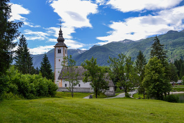 Old stone church on the shore of a mountain lake. Green mountain range and blue sky with white clouds.
