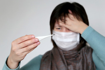 Fever and coronavirus symptoms, woman with a scarf wrapped around her throat measures body temperature. Worried girl in medical mask looks at digital thermometer in her hands, concept of cold and flu