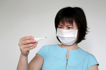 Woman in medical mask measures body temperature. Worried girl looks at digital thermometer in her...