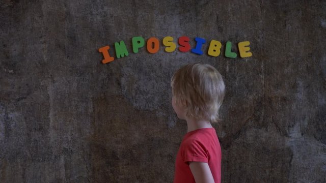 funny blond boy in red clothes is standing against wall where word is written: impossible. Boy does not like this word and he covers first two letters with his palms in order to succeed: possible