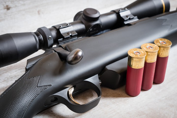 A bolt action scoped shotgun and three shotgun shells against a light wooden background. The...