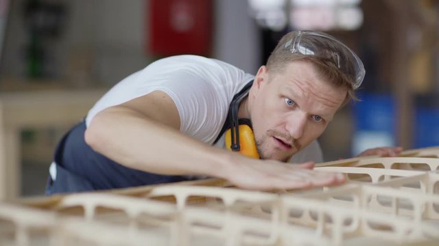 Rack focus of middle aged carpenter touching carefully unfinished wooden boat model placed on workbench checking if wood is smooth enough