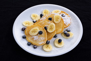 pancakes with banana and blueberries on a white plate, tasty and beautiful on a black background