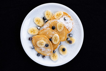 pancakes with banana and blueberries on a white plate, tasty and beautiful on a black background
