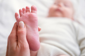 Massage. Close up baby feet in mother hands. Copyspace. Young caucasian woman makes massage for happy infant baby on white bed at home