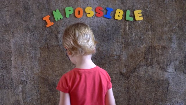 funny blond boy in red clothes is standing against wall where word is written: impossible. Boy does not like this word and he covers first two letters with his palms in order to succeed: possible