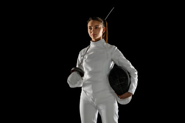 Confident. Teen girl in fencing costume with sword in hand isolated on black background. Young female model practicing and training in motion, action. Copyspace. Sport, youth, healthy lifestyle.