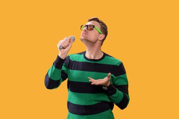 Young man in glasses singing song in microphone. Young posing isolated on yellow orange background. People lifestyle concept.