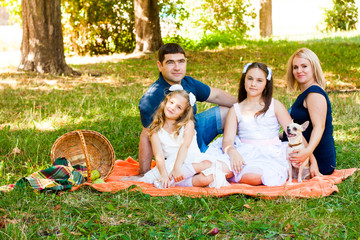 Happy Two Parent Family With Two Daughters And Dog On Orange Picnic Blanket In The Park