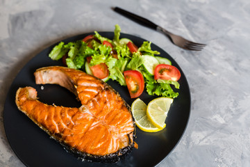 Fried salmon fish steak with vegetable salad and lemon on black plate. Creative layout made of fish, lettuce, tomato, cucumber, top view, flat lay, mockup, overhead. Healthy food concept