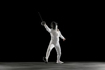 Fototapeta na wymiar Targets. Teen girl in fencing costume with sword in hand isolated on black background. Young female model practicing and training in motion, action. Copyspace. Sport, youth, healthy lifestyle.