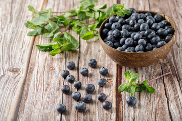 Fresh blueberries in a coconut bowl with mint sprigs are scattered on an rustic brown wooden table 