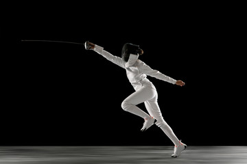 Obraz premium Catching moment. Teen girl in fencing costume with sword in hand isolated on black background. Young female model practicing and training in motion, action. Copyspace. Sport, youth, healthy lifestyle.