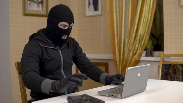 A robber is trying to hack into a laptop. A masked thug is sitting in a house and trying to break into a laptop. Theft of data from a computer.