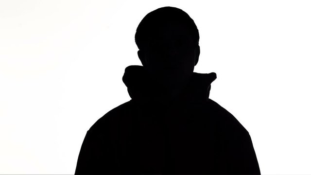 silhouette of a young man puts on a hood of a sweatshirt and goes on white isolated background, unknown person, guy hides face, concept fashion