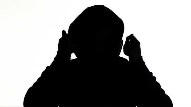silhouette of a young man puts on a hood of a sweatshirt and goes on white isolated background, unknown person, guy hides face, concept fashion