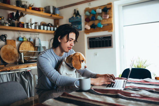 young man holding his dog in nap while using laptop in kitchen