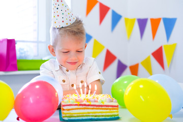 Fototapeta na wymiar Blonde caucasian boy sits thoughtfully and dreamily at festive table near birthday rainbow cake and makes a wish. Colorful background with balloons.