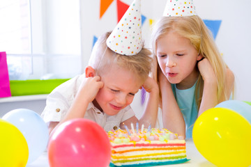 Fototapeta na wymiar Two blonde caucasian kids boy and girl have fun blowing out candles at birthday rainbow cake with burning candles at birthday party