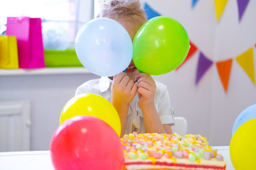 Fototapeta na wymiar Birthday boy hid behind colorful balloons near birthday rainbow cake. Festivel background. Funny birthday party.