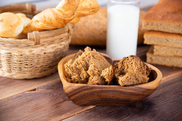 Broken bread in pieces in a wooden plate and other types of bakery products with a glass of milk on a dark table