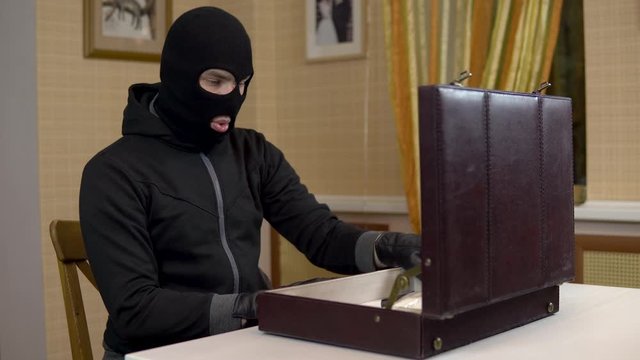 The robber opened the suitcase. A masked thug is sitting in a house, taking drugs and money from a suitcase.