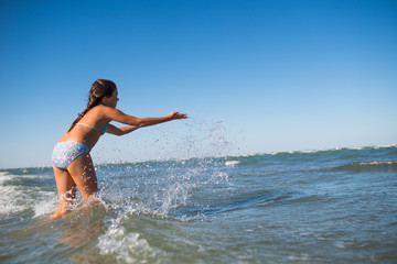 Fototapeta premium Little funny active happy girl splashing in the noisy sea waves on a sunny warm summer day. Sea vacation concept with kids. Advertising space