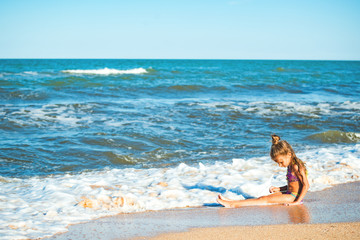 Fototapeta na wymiar Little positive girl sits on the seashore and enjoys the sea waves on a sunny summer day during vacation. Concept of family holidays with children
