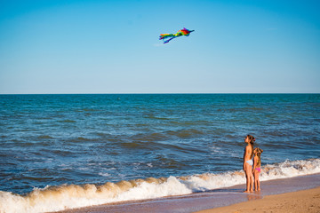 Two cheerful little girls sisters play with a kite running on the sandy shore by the sea on a sunny warm summer day. Concept of children's games and active entertainment. Advertising space