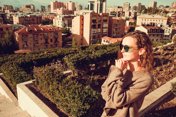 Stylish young caucasian woman in sweater enjoy view from cascade complex. Yerevan cityscape. City tour. Girl travel to Armenia. Tourism concept. Sunny autumn day. Armenian architecture. Green bushes