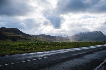 Iceland's ring road lit up by beautiful rays of sunshine making their way through a cloudy sky. Snow capped peaks in the background and animals and lush greenery makeup the midground