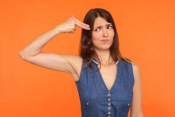 Desperate brunette woman in denim dress looking frightened at finger gun pointed to head, killing herself with suicide gesture, scared, tired of life. indoor studio shot isolated on orange background