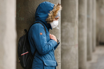 Unhealthy man with a hood holds hands on chest, wearing protective facial mask against transmissible infectious diseases and as protection against the flu or coronavirus in public places, outdoor.