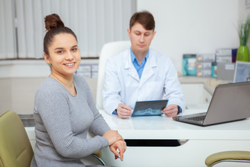 Happy healthy young woman smiling to the camera, her doctor examining x-ray scan on the background