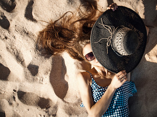 Beautiful young girl in sunglasses and a hat in a blue swimsuit on a golden beach