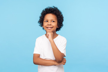 Portrait of thoughtful happy little boy in T-shirt standing with pensive look and touching his...