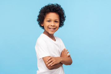 Portrait of happy and confident little boy in white T-shirt standing with crossed hands and smiling at camera, healthy cheerful child, positive emotions. indoor studio shot isolated on blue background © khosrork