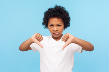 Dislike! Portrait of dissatisfied upset little boy with curly hair showing thumbs down gesture and looking displeased, naughty child making disagree disapprove finger sign. studio shot blue background - obrazy, fototapety, plakaty